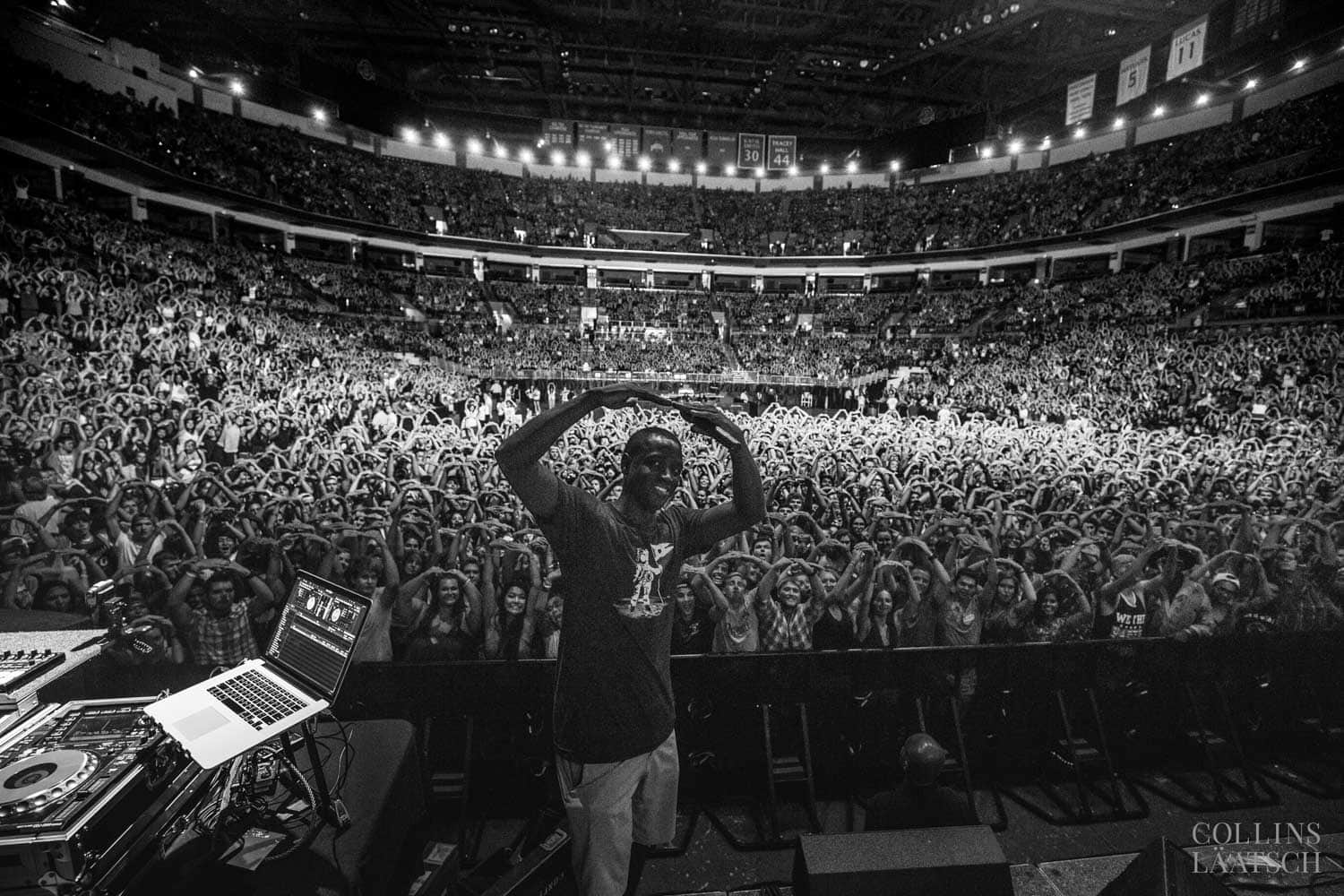 A performer on stage faces a large, cheering audience, with a laptop and DJ equipment in the foreground.