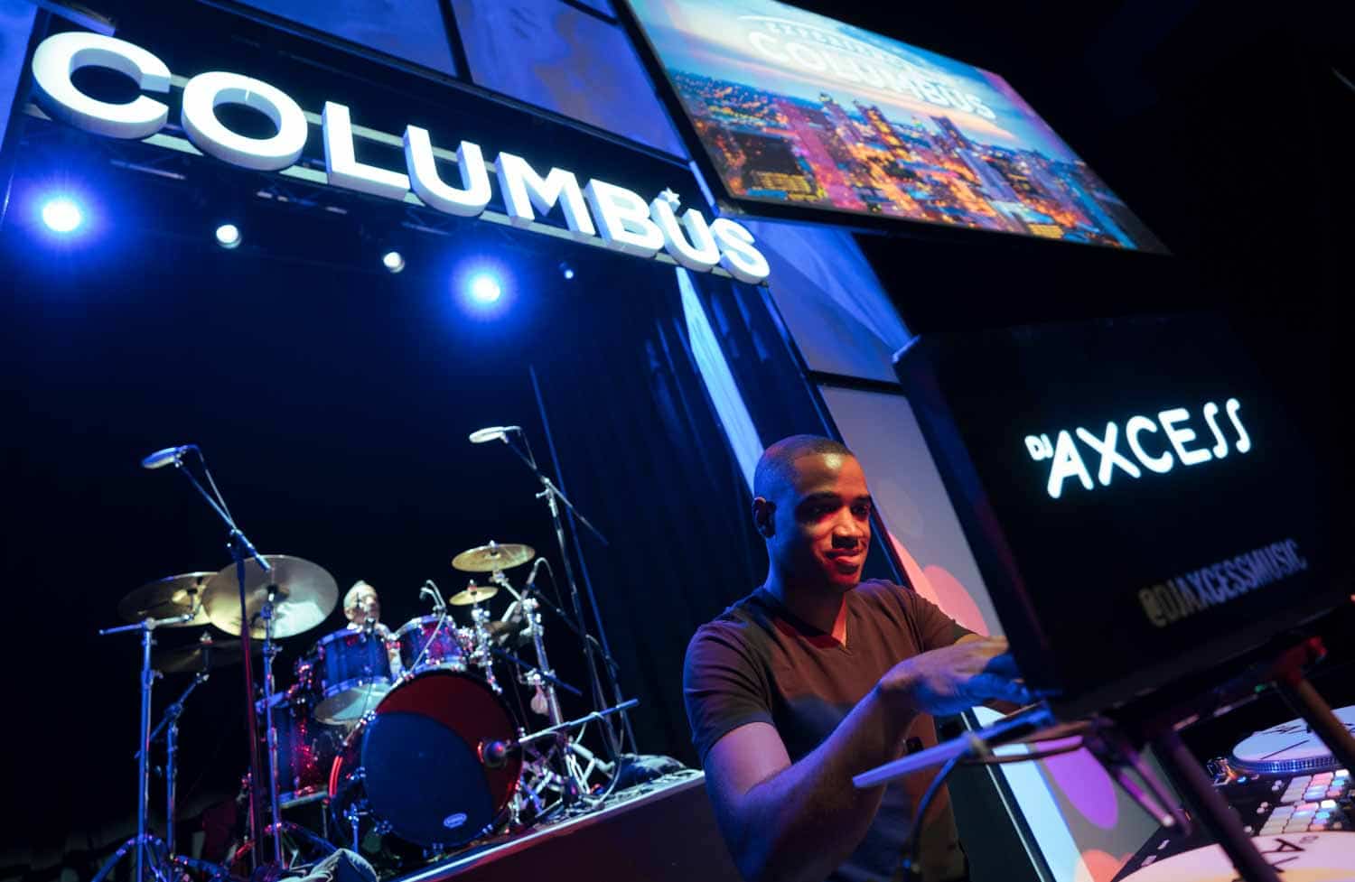 DJ performing on stage with a drummer in the background, under a sign that reads Columbus.