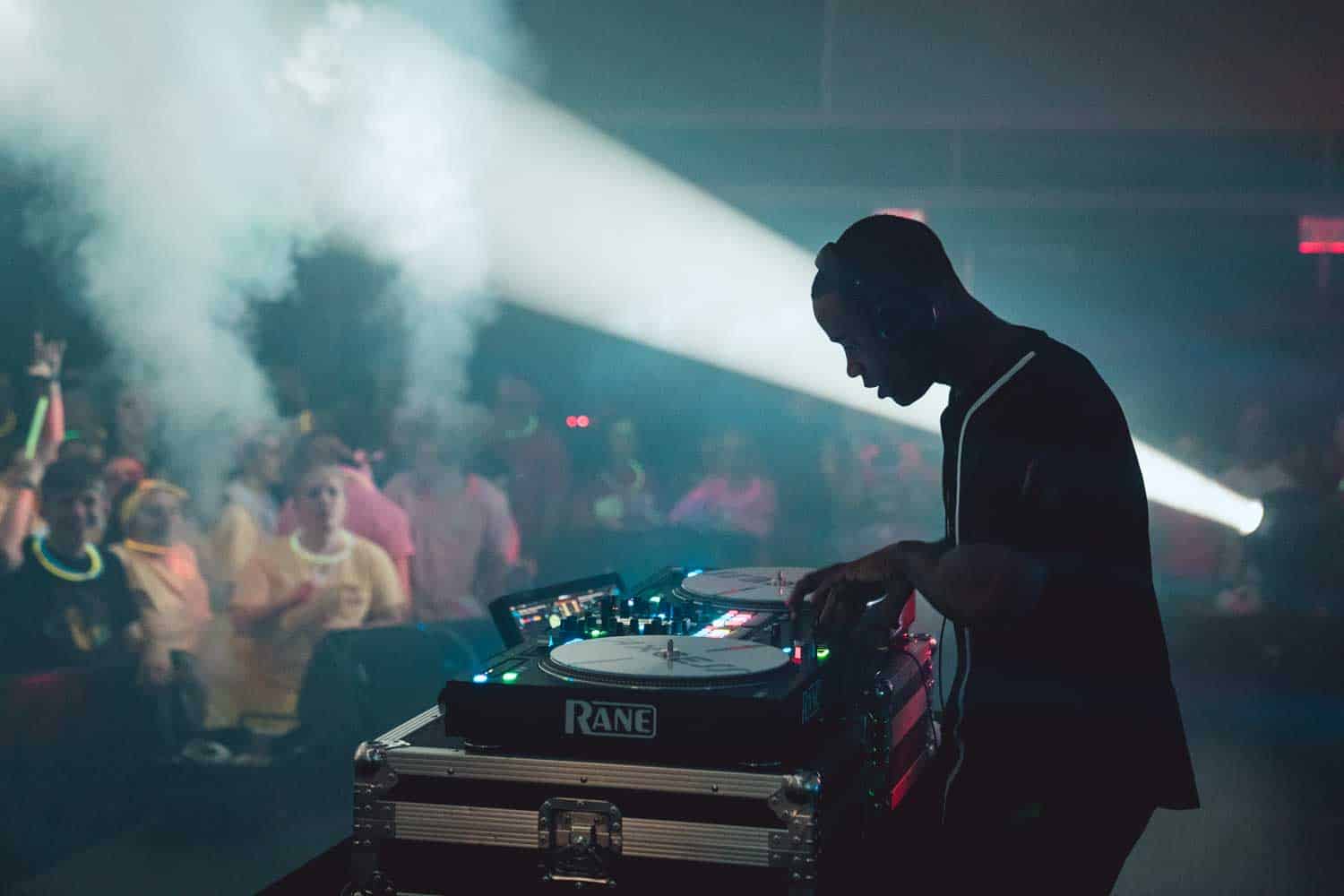 A DJ is performing on stage with a crowd dancing in the background. Bright lights and smoke create an energetic atmosphere.