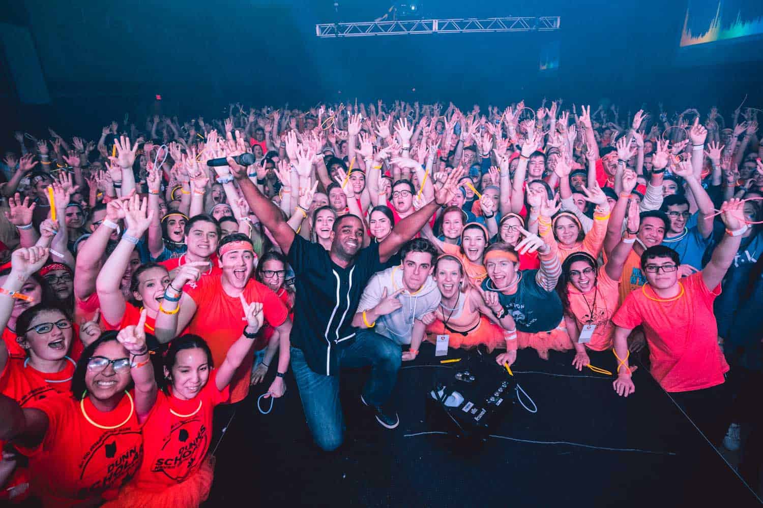 A large group of people in colorful shirts pose energetically for a photo at an indoor event, with raised hands and smiling faces.