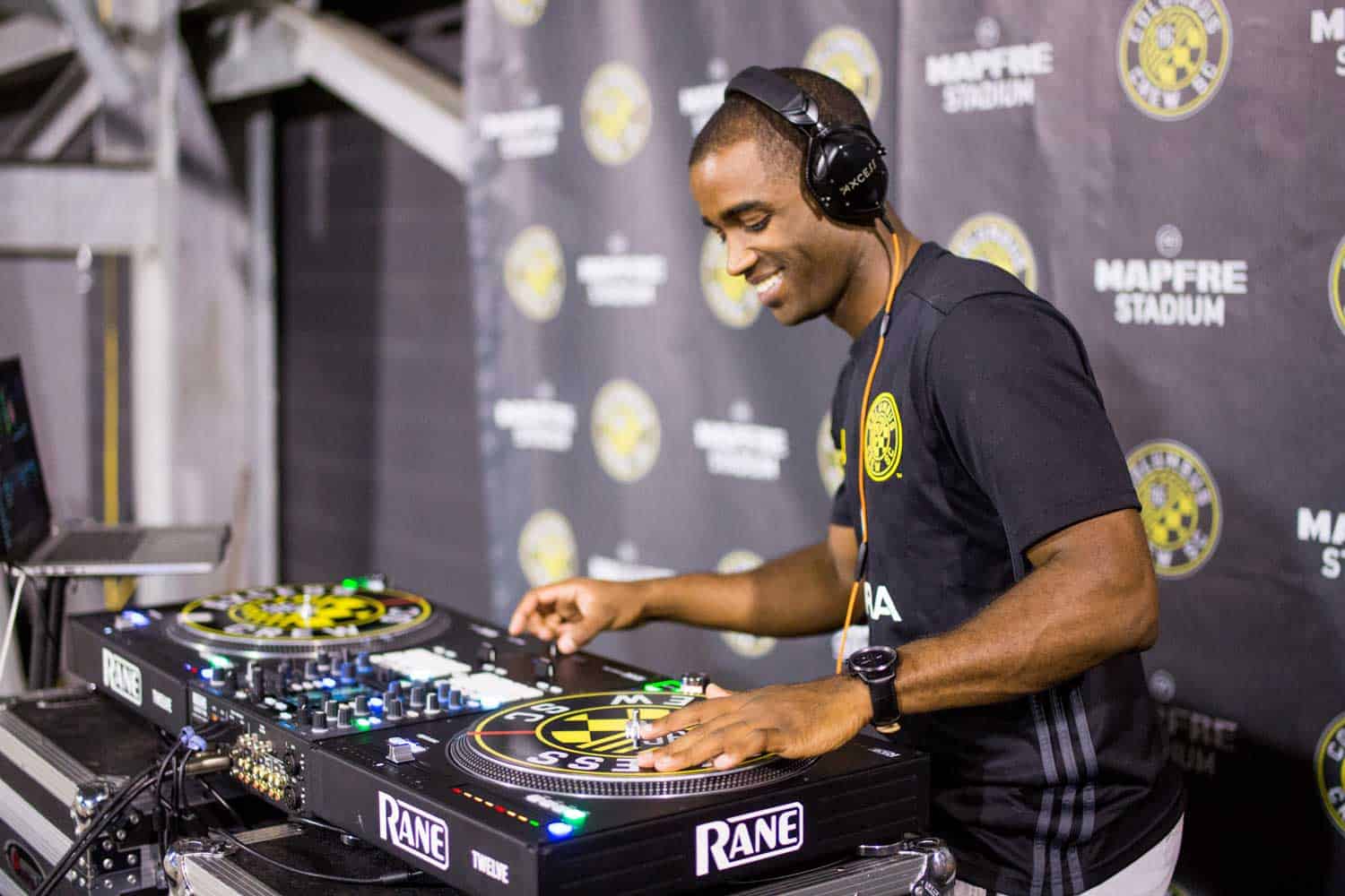 DJ wearing headphones mixes music on turntables at a venue with a Mapfre Stadium backdrop.