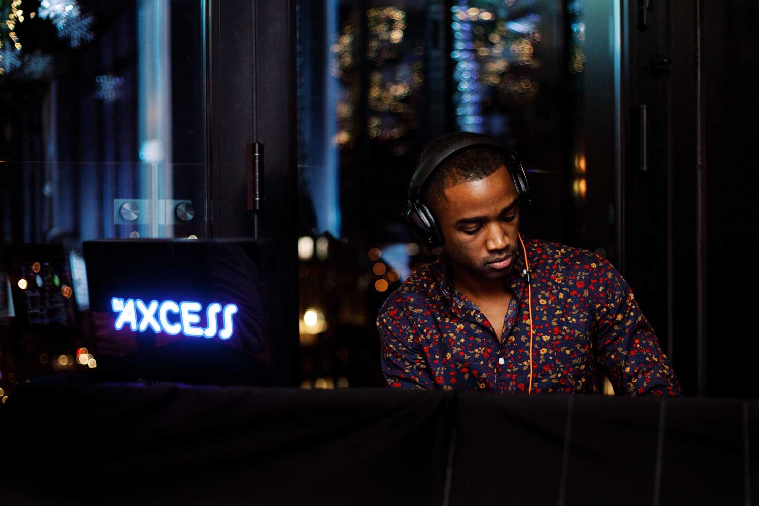 A man wearing headphones and a patterned shirt is DJing at a nightclub with a AXCESS sign in the background.