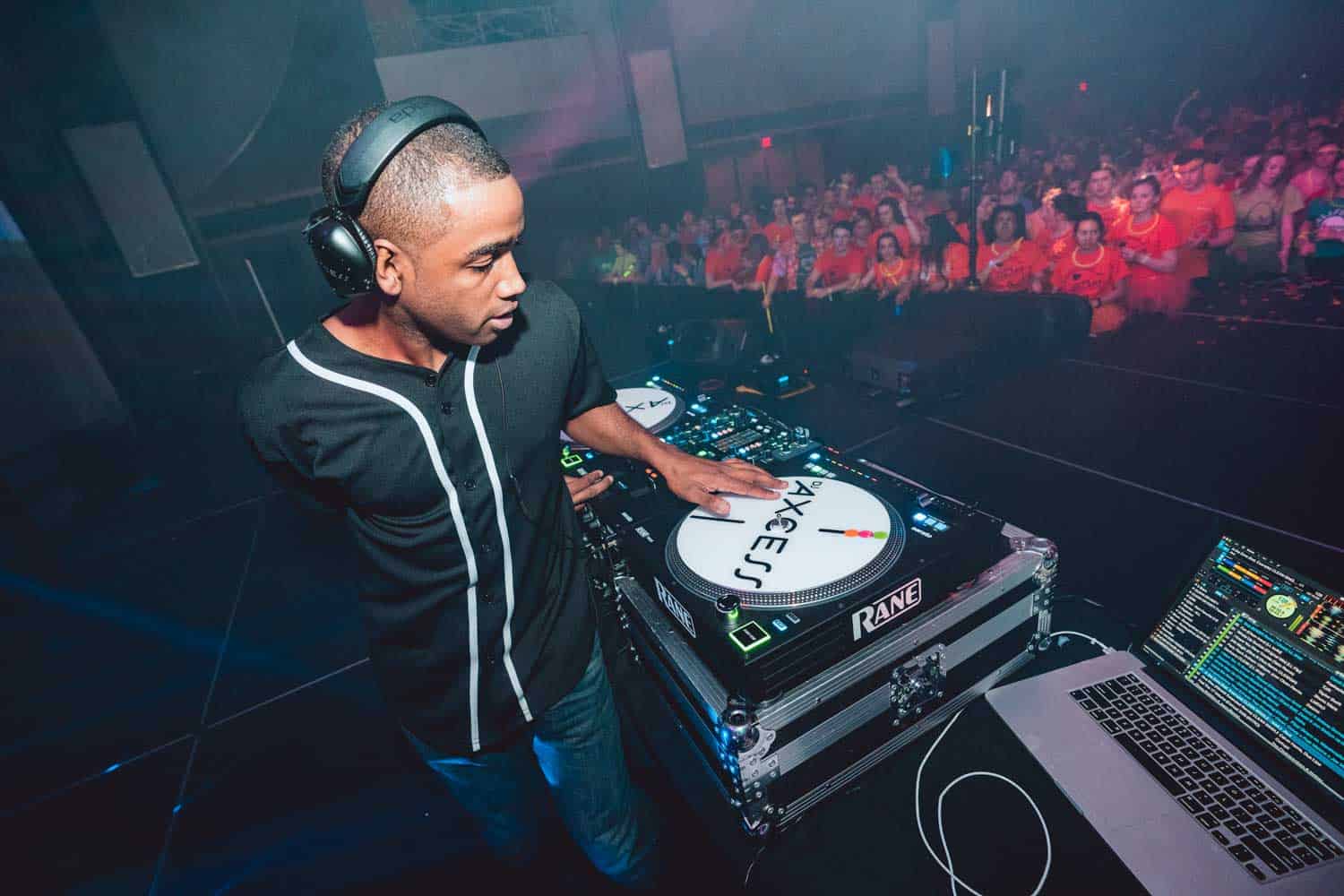 DJ performing on stage, using turntables and a laptop, with a crowd watching in the background.