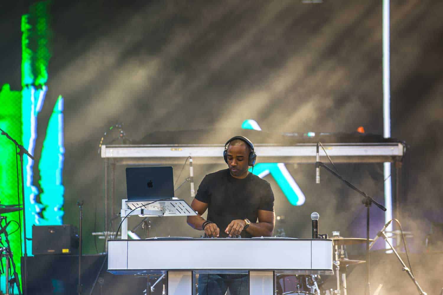 A DJ wearing headphones is performing on a stage with equipment, including a laptop and sound mixer, surrounded by mist and light beams.