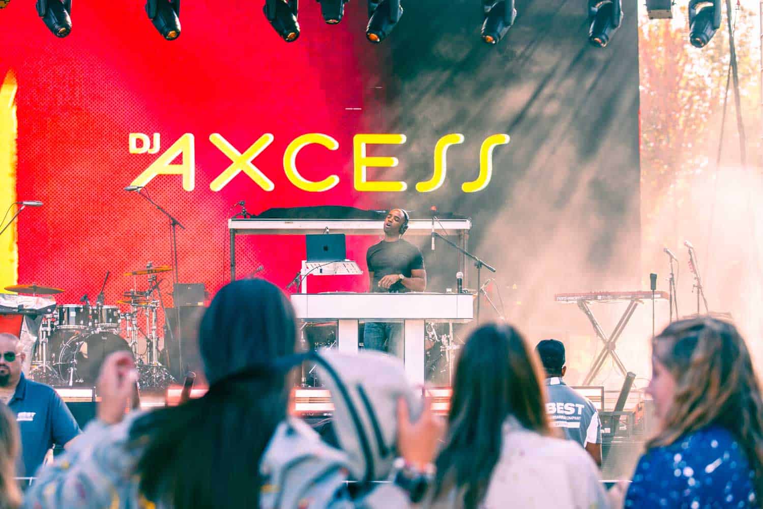 DJ performing on stage with a bright AXCESS sign in the background, surrounded by musical instruments and lighting, while an audience watches.