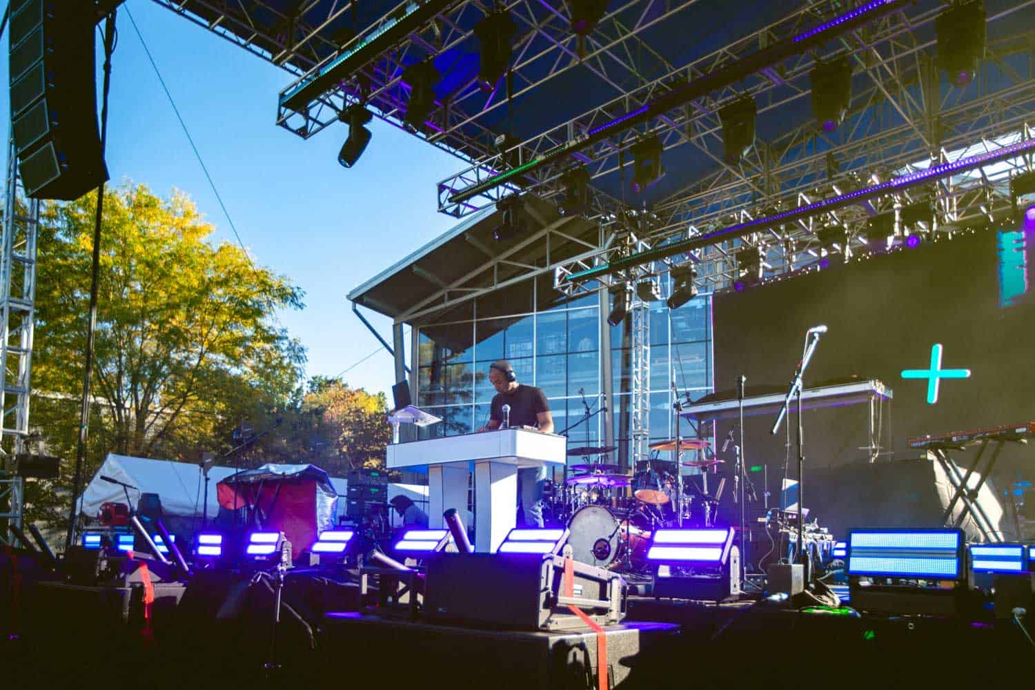 A man stands at a DJ booth on an outdoor stage with colorful lights and a large screen in the background. Trees and a building are visible under a clear sky.