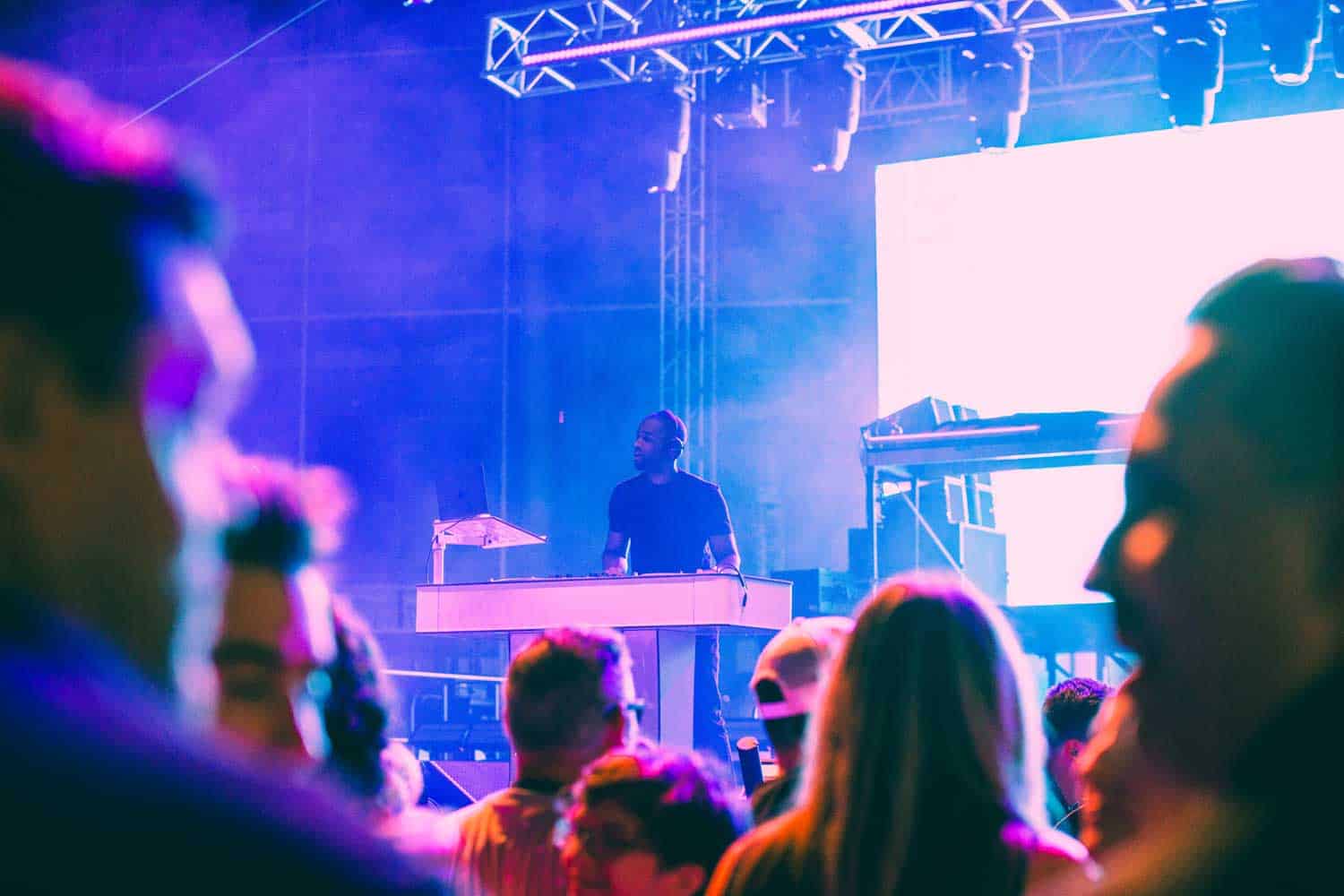 A DJ performs on stage with colorful lighting effects, surrounded by a crowd of people at a concert.