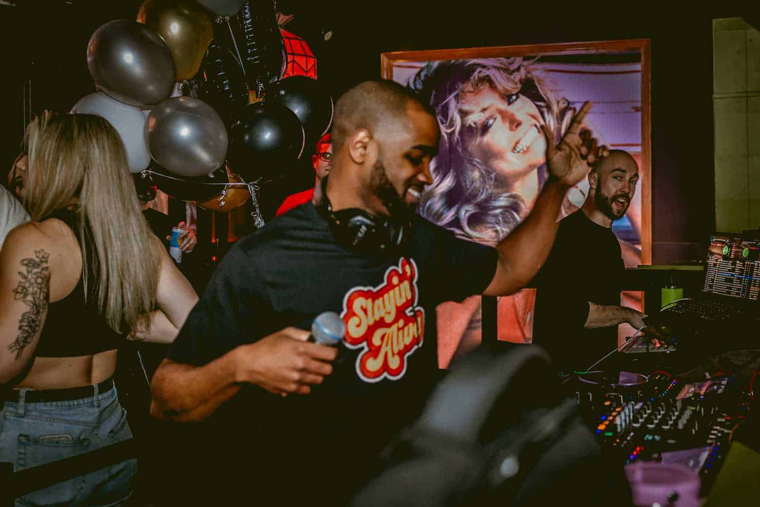 A DJ wearing headphones holds a microphone in a lively club. Balloons and a large poster of a smiling woman are in the background.