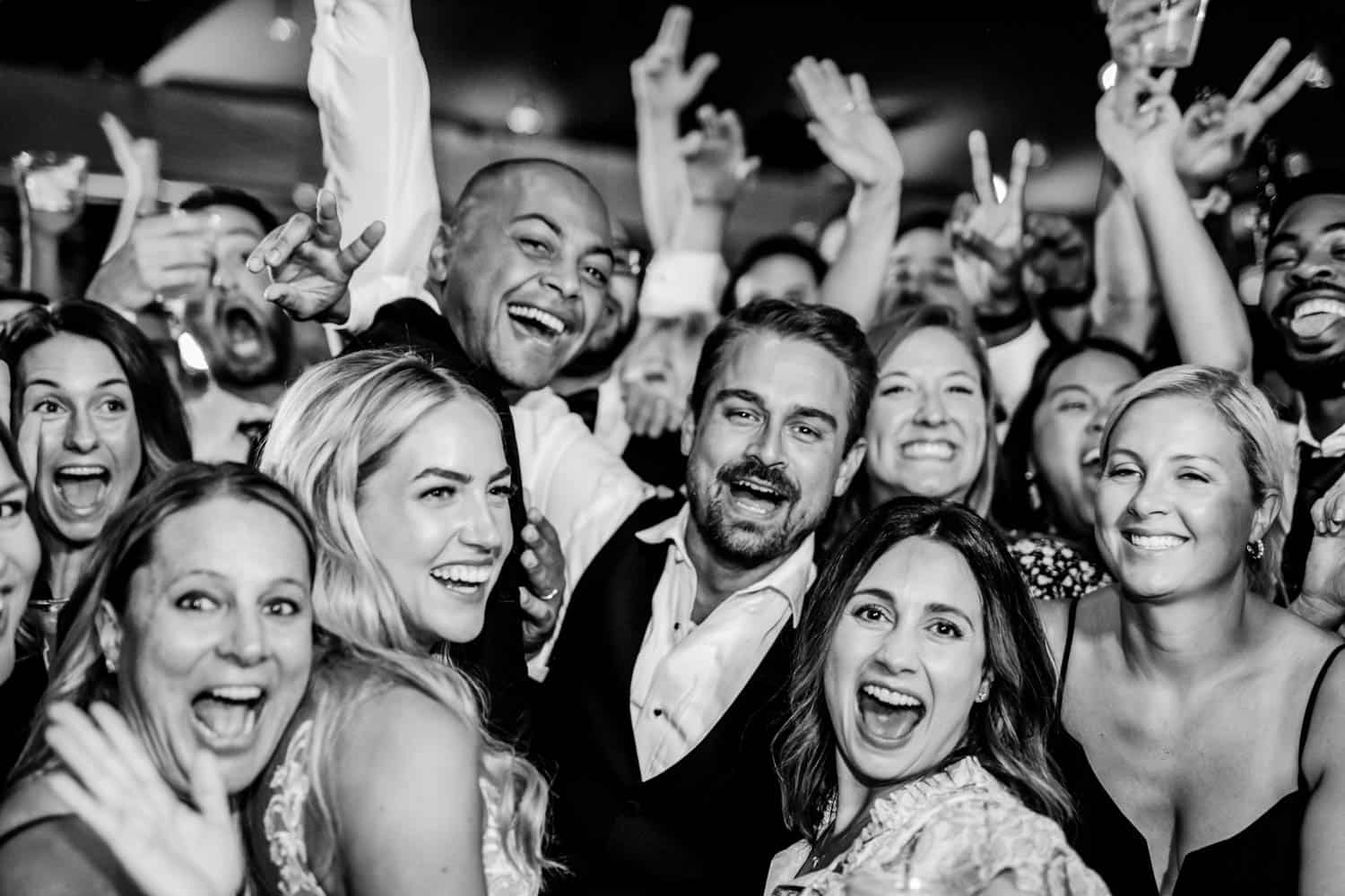 A group of people smiling and posing for a photo at a crowded event, with many making peace signs. The image is in black and white.