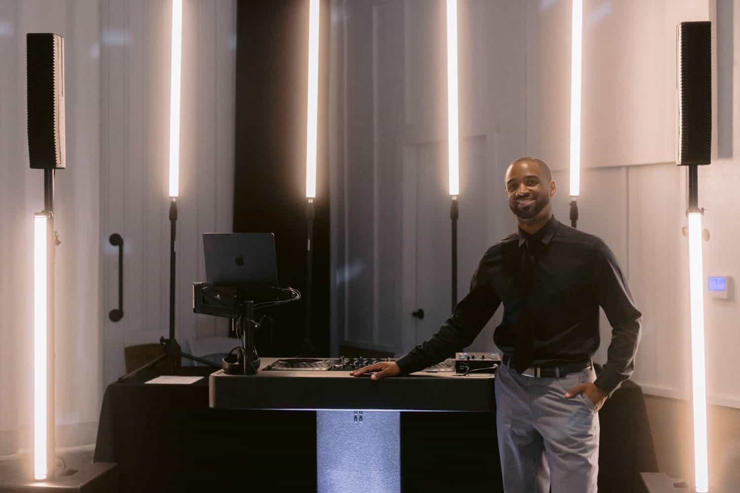 A man in formal attire stands beside DJ equipment, surrounded by vertical lights in a dimly lit room.