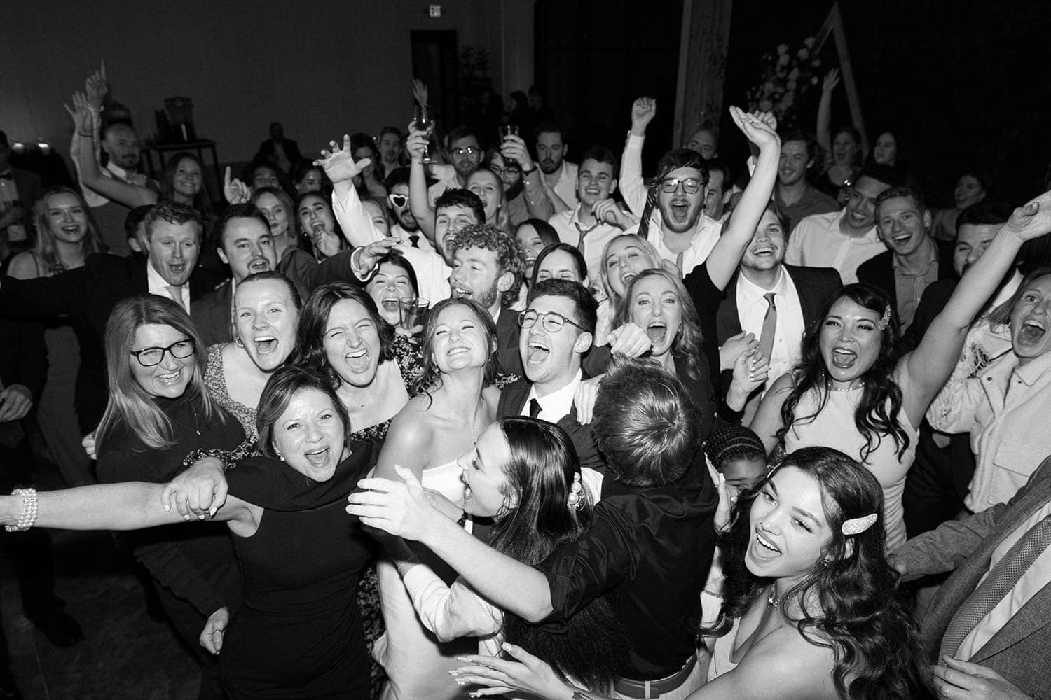 A large group of people, dressed in formal attire, are gathered closely together, smiling and raising their arms in celebration at an indoor event.