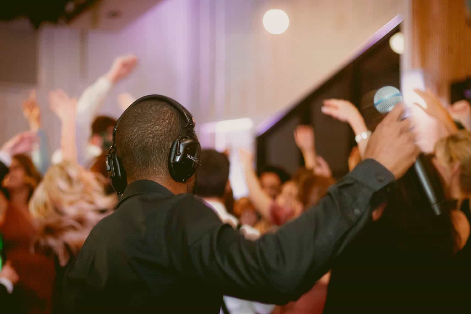 A DJ wearing headphones holds a microphone and faces a lively crowd with raised hands in a dimly lit venue.