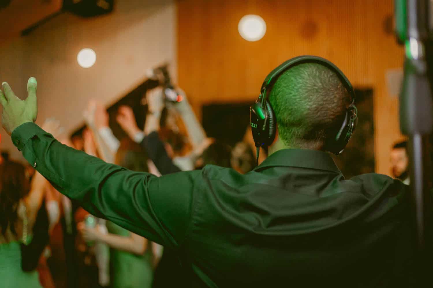 Man wearing headphones with arms raised, facing a crowd of people dancing in a dimly lit room.