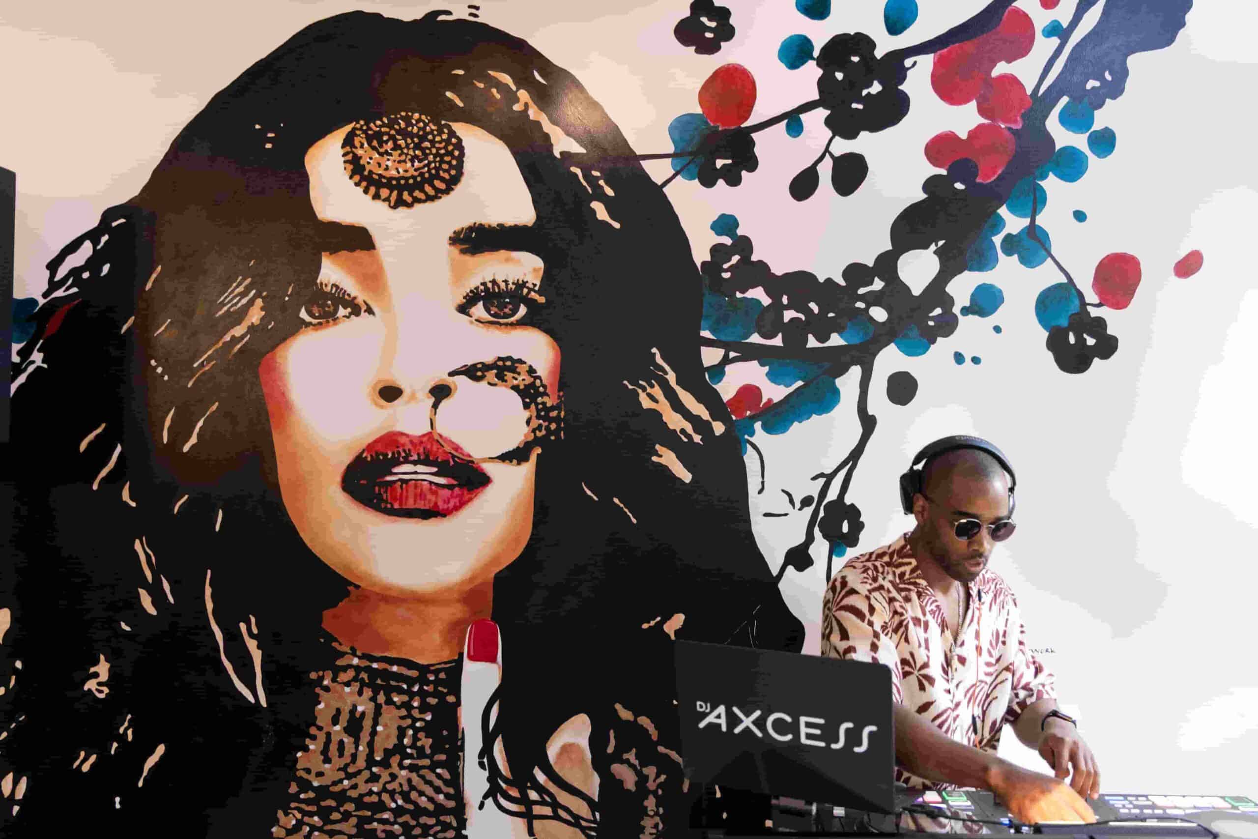 DJ AXCESS performing on a controller in front of a vibrant mural featuring a stylized woman’s face with bold makeup and abstract paint splashes, creating a modern, artistic event backdrop.