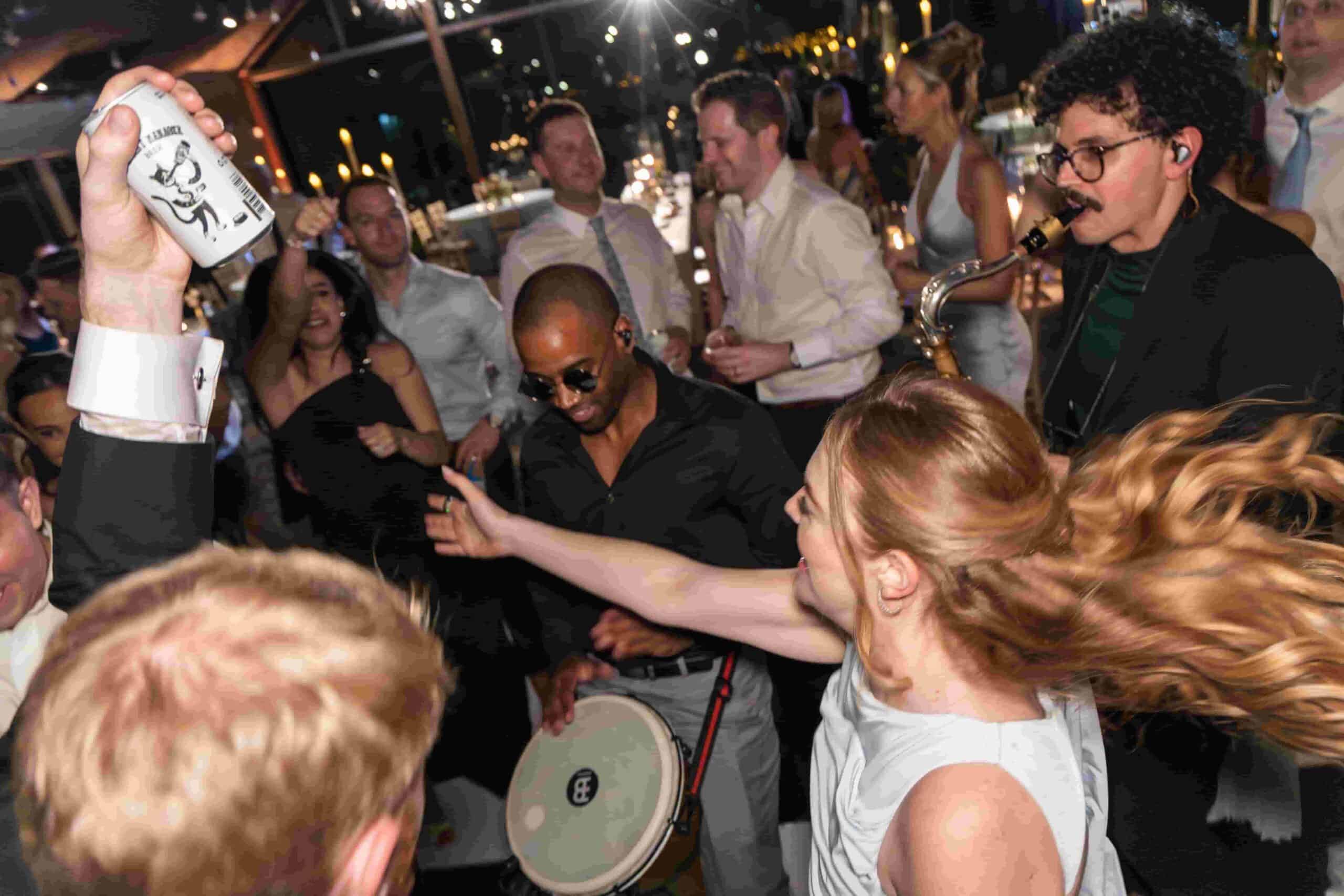 DJ AXCESS performing live among a packed dance floor alongside a saxophonist and percussionist, with guests dancing, raising drinks, and interacting closely in a high-energy wedding or event setting.