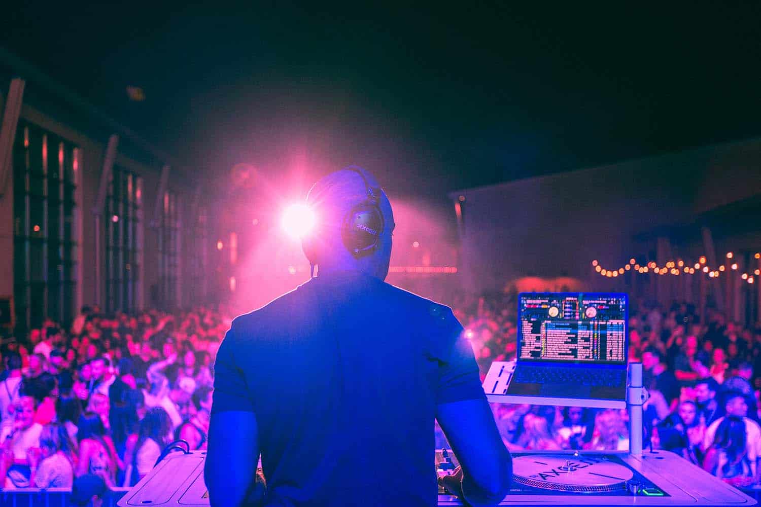 DJ axcess performing from behind the booth in front of a large, packed crowd at night, with bright stage lighting, abercrombie the challenge, high-energy party.