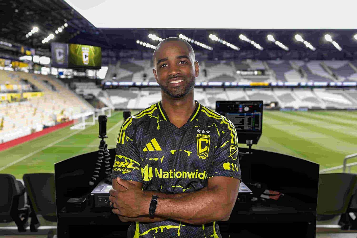 DJ AXCESS standing in front of a DJ setup inside a large soccer stadium, wearing a Columbus Crew jersey with arms crossed and the field visible in the background.